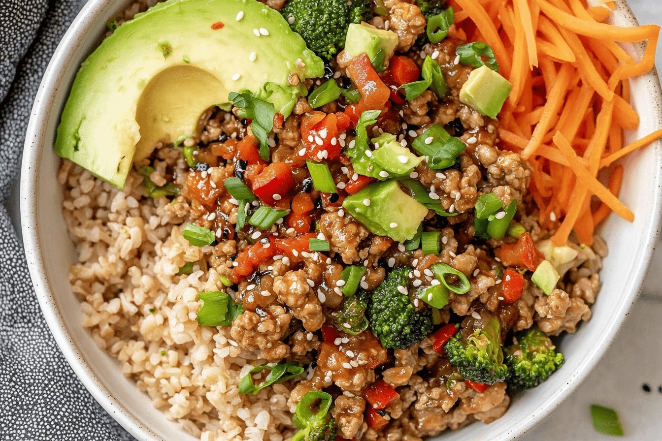 Overhead close-up of a Teriyaki Turkey Rice Bowl with avocado slices, shredded carrots, broccoli, green onions, and sesame seeds on brown rice.